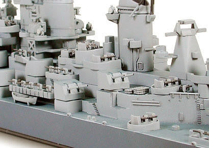 A close-up of a light gray unpainted model ship or naval vessel, showing detailed deck structures, turrets, and equipment in a studio setting.